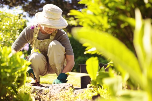 A woman working in a well-maintained garden during daylight hours, crouched down near the ground and wearing a wide-brimmed hat, beige overalls, and gardening gloves. She appears to be tending to flower beds or small shrubs, with various garden tools visible nearby. The garden features a lush, green lawn with neatly edged flower beds, a mixture of vibrant flowering plants, and leafy shrubbery. The background shows mature trees providing shade, and the scene is illuminated by bright, natural sunlight indicating clear weather. The overall environment suggests a peaceful outdoor space, possibly in Sydenham, with a focus on tending, landscaping, and gardening activities that include soil care and plant maintenance. This setting aligns with gardening services offered by Landscapers Sydenham, highlighting outdoor garden upkeep and landscaping work, especially during the growing season.