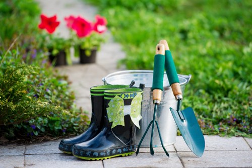 The image depicts a section of an outdoor garden area with a paved pathway and a well-maintained grassy lawn beyond. In the foreground, there are a pair of black rubber gardening boots with a green and black striped fabric cuff, placed on the stone surface. Next to the boots, there are gardening tools including a small trowel with a blue handle and a hand rake with a green handle, leaning against a white plastic bucket. The background features a lush, green garden border with dense shrubbery and flowering plants, including a few potted plants with pink blooms. The overall scene suggests a tidy and organized outdoor space prepared for planting or garden maintenance, typical of professional gardening and landscaping services in Sydenham, with natural lighting indicating a mild, sunny day. The image emphasizes outdoor gardening equipment and a landscaped garden environment consistent with services provided by Landscapers Sydenham.