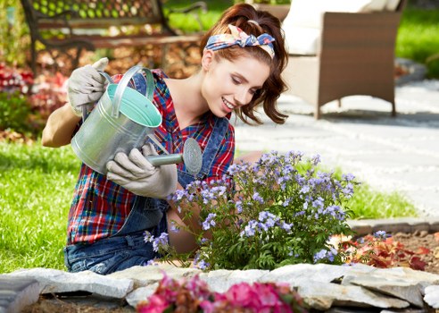 A young woman with light skin, wearing a plaid shirt, denim overalls, gloves, and a patterned headband, is tending to a flower bed in a well-maintained garden. She is smiling while carefully watering blooming purple and pink flowers with a metal watering can. The garden features a neatly edged flower bed bordered with stone and filled with vibrant flowering plants, surrounded by lush green grass. In the background, there is outdoor patio furniture including a table and chairs, indicating a landscaped yard in Sydenham, South London, with sunlight illuminating the scene and clear weather. The overall setting reflects a well-kept outdoor space suitable for gardening and lawn care services provided by Landscapers Sydenham, emphasizing plant cultivation, garden maintenance, and outdoor aesthetic enhancement.