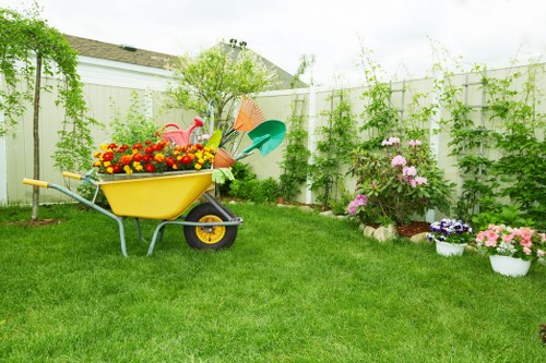 A lush, well-maintained backyard garden in Sydenham featuring a vibrant green lawn with freshly cut grass, bordered by a tall white fence adorned with climbing plants and greenery. In the foreground, a yellow wheelbarrow filled with colourful flower pots and gardening tools such as a small green watering can, an orange spade, and a blue watering can rests on the grass. To the right, there is a flower bed with blooming pink, purple, and white flowers, bordered by a narrow strip of soil. In the background, a small shrub or young tree adds height and structure to the garden, while the overcast sky provides diffused natural lighting. This outdoor space demonstrates a tidy, inviting garden ideal for landscaping and outdoor maintenance, reflecting the expertise of Landscapers Sydenham in garden care and fencing enhancements.
