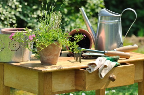 A wooden outdoor table situated in a garden with various gardening tools and supplies arranged on its surface. Visible elements include potted plants with green foliage, some with flowering pink and purple blooms, indicating healthy plant growth. A metal watering can with a curved handle and a spout is placed on the table, alongside hand tools such as a small garden fork with metal tines, pruning shears, and a trowel with a wooden handle. A pair of gardening gloves, made of fabric with green accents, rest draped over the edge of the table. The background features lush green trees and shrubs, suggesting a well-maintained garden environment. The scene depicts typical outdoor gardening activities, with a focus on plant care and garden maintenance, reflective of professional gardening services such as those offered by Landscapers Sydenham, in a natural, sunlit setting in Sydenham or nearby areas, with soft lighting and a clear, pleasant weather condition. The table's wooden surface displays a natural texture, complementing the outdoor atmosphere.