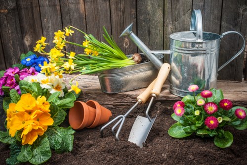 A garden scene featuring freshly turned dark soil prepared for planting, with a background of weathered wooden fencing. In the foreground, there are vibrant flowering plants including yellow primroses, pink primulas, and blue-flowered plants, along with empty terracotta plant pots. Gardening tools such as a small hand rake, trowel, and a watering can are arranged on or near the soil, indicating ongoing planting or garden maintenance activities. The image captures a well-maintained outdoor space typical of residential gardens in Sydenham, highlighting the readiness for planting and landscaping work that a professional gardening service like Landscapers Sydenham may offer for nearby homes and properties.