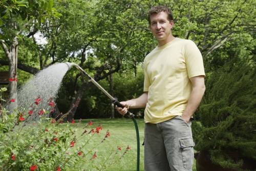 A man wearing a pale yellow t-shirt and grey trousers is standing in a well-maintained garden, holding a garden hose and watering the plants and flower beds. The garden features a lush green lawn with neatly trimmed grass, bordered by a variety of shrubs and flowering plants, including red blooms. In the background, there are tall trees with dense foliage providing shade, along with a curved hedge that adds structure to the outdoor space. The scene is outdoors on a bright day, with natural light illuminating the vibrant greenery and colorful flowers, creating a peaceful and inviting environment typical of landscaped gardens in Sydenham. This setting supports outdoor gardening and lawn care services offered by companies like landscaperssydenham.co.uk, ensuring a tidy and flourishing garden appearance.