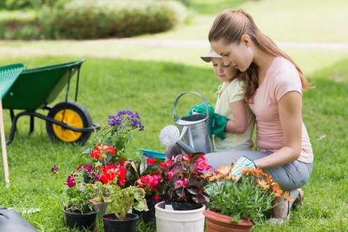 A woman and a young girl are kneeling on a lush, well-maintained lawn in a garden, engaged in gardening activities. They are surrounded by a variety of potted flowers, including red, purple, and orange blooms, arranged in a semi-circle in front of them. The woman is wearing a pale pink T-shirt and jeans, with her hair tied back, while the girl is dressed in a yellow T-shirt and gloves, holding a small watering can. In the background, a green garden wheelbarrow with yellow wheels is visible on the grass, and a neatly trimmed hedge runs along the edge of the garden, with some trees further back under clear, bright weather conditions. The garden appears spacious, with a mixture of flowering plants and open lawn area, ideal for outdoor gardening and family activities, illustrating a well-kept outdoor space suitable for professional gardening and landscaping care by companies like Landscapers Sydenham.