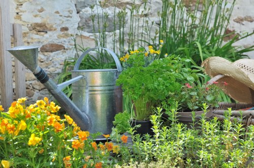 A lush garden scene featuring a variety of plants and flowers, with a foreground of vibrant yellow and orange flowering plants. Behind these, there are green leafy shrubs and smaller herbs, arranged in a natural, slightly informal layout. To the right, a wooden gardening hat rests on a garden surface, and in the center, a silver metal watering can is placed among the greenery, suggesting ongoing garden maintenance. The garden is set against a stone wall, adding rustic charm, and taller plants with thin stems and yellow blooms grow towards the back, providing height contrast. The overall environment appears well-tended, with natural light illuminating the scene, enhancing the lush green tones and colorful blossoms, reflecting typical gardening activities undertaken by a professional landscapers Sydenham service in the local area.