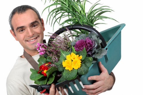 A man with short dark hair and a friendly expression is holding a light blue plastic garden watering can filled with a variety of colourful plants, including purple, yellow, and red flowers, along with green foliage. The background is plain white, emphasizing the garden elements and the man's light grey clothing. The scene reflects outdoor gardening activities, with the plants suggesting healthy growth and vibrant planting efforts typical of a well-maintained garden in Sydenham or broader South London areas. The watering can appears to be used for watering or establishing garden flowers and plants, underscoring services related to planting, watering, or garden care. The image's lighting is bright and even, creating a cheerful and professional atmosphere suitable for a landscaping or gardening website like landscaperssydenham.co.uk, which emphasizes quality outdoor maintenance and plant care in the local area.
