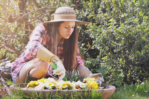 A young woman wearing a wide-brimmed straw hat, beige gardening gloves, and a plaid shirt is kneeling on a lush green lawn in a backyard garden. She is carefully planting or tending to a flower bed that contains a variety of colourful blooming flowers, including yellow, white, and purple blossoms. The garden features well-maintained grass, neatly bordered flower beds, and dense shrubbery and trees in the background, providing shade and natural privacy. The scene is illuminated by natural daylight, suggesting a clear, mild weather day suitable for outdoor gardening. This setting exemplifies typical gardening activities that a professional landscaper like Landscapers Sydenham might undertake, focusing on garden care, plant maintenance, and outdoor aesthetics in residential outdoor spaces near Sydenham, London, and surrounding areas.